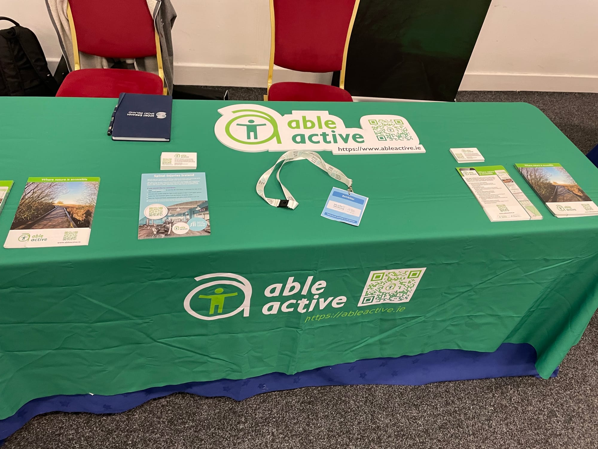 Exhibitor table featuring Able Active branded green tablecloth with flyers, exhibitor pass and business cards.