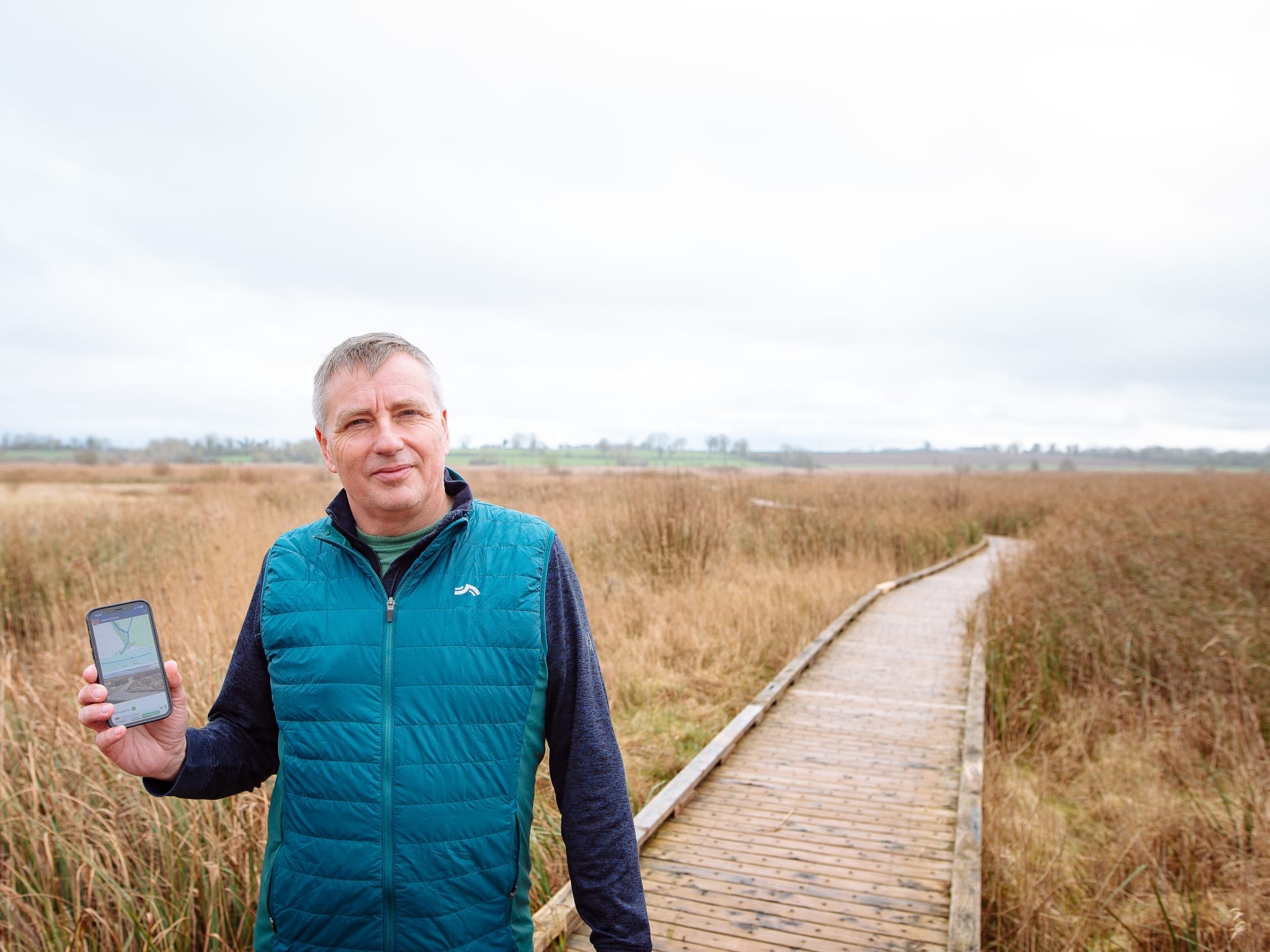 Outdoor photo of Adrian Geissel holding a mobile phone showing the Able Active app while standing on a wooden boardwalk